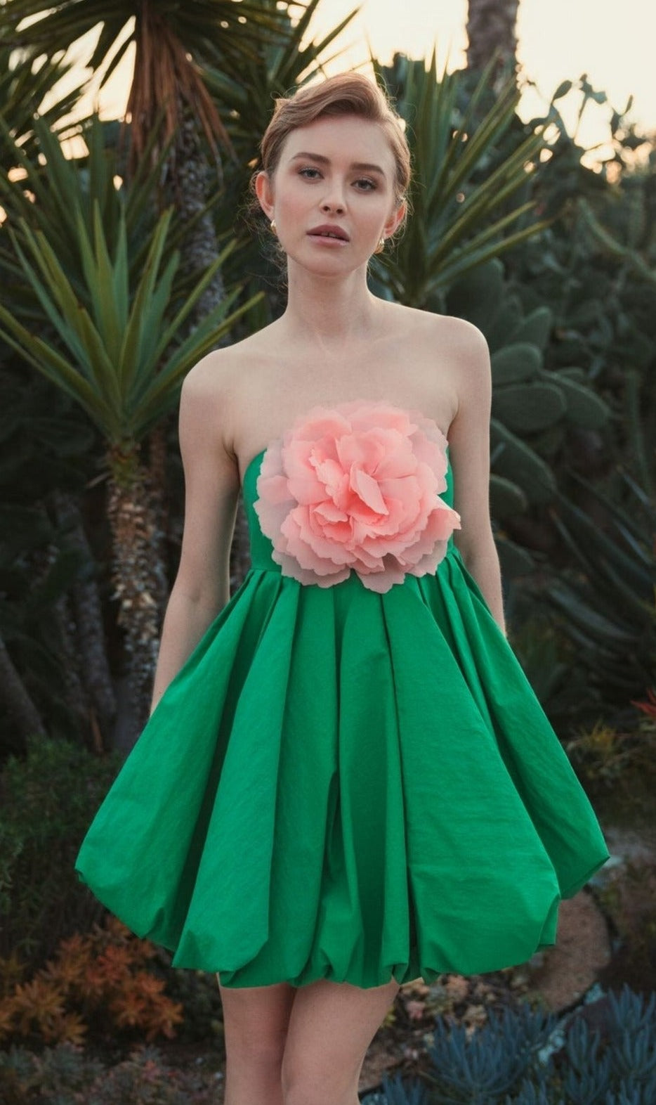 Outdoors among lush plants and cacti, a woman wears the Mac Duggal 20931 Short Bubble Party Dress, strapless and adorned with a large pink flower on the bodice.